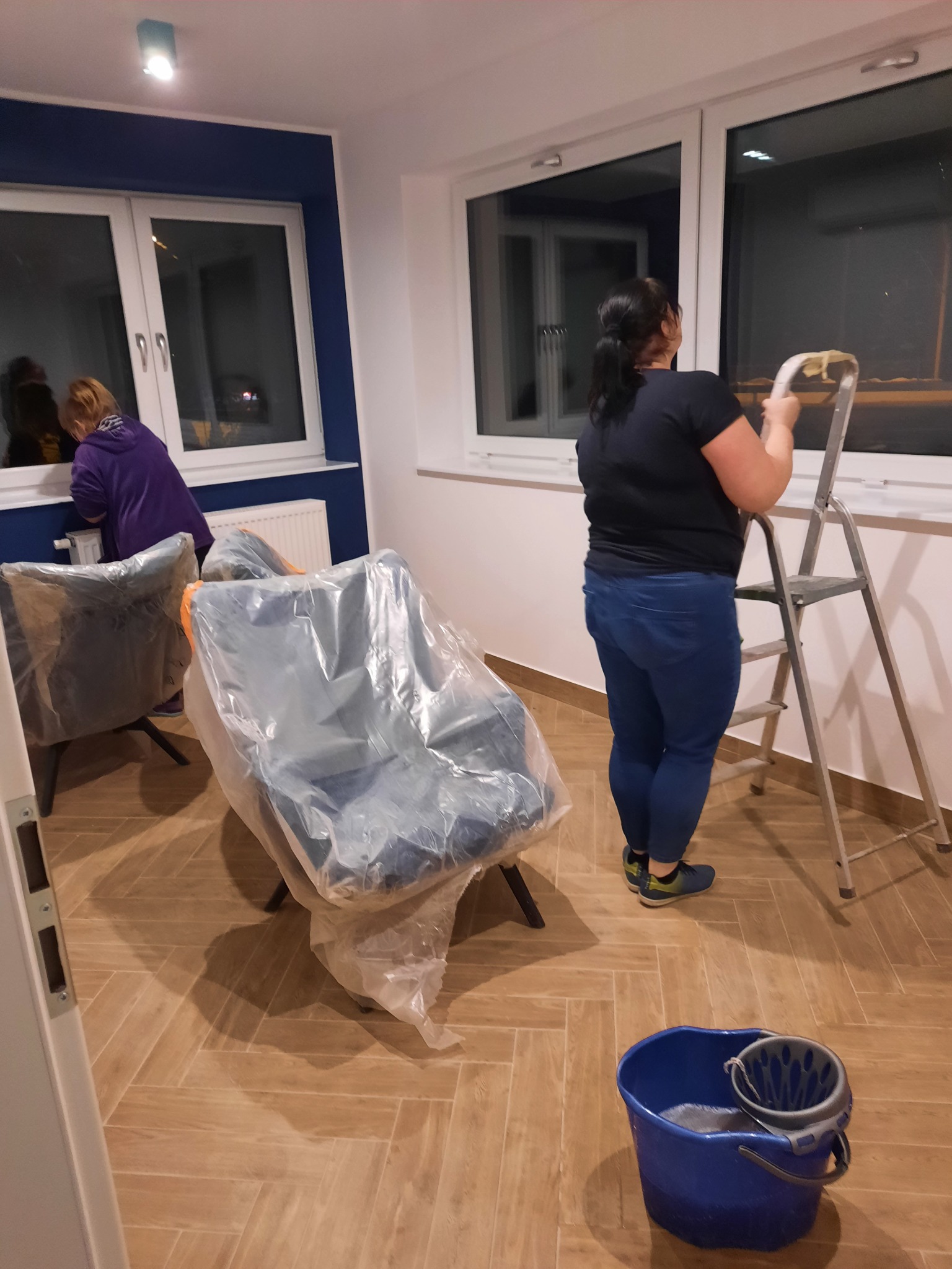 Kobieta na drabinie myje okno w nowym mieszkaniu, krzesła zabezpieczone folią, widoczne wiadro z wodą, druga osoba czyści okno obok.