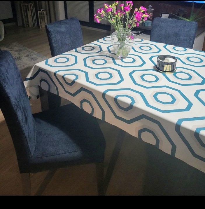 Stół nakryty wzorzystym obrusem w geometryczne kształty, na nim wazon z różowymi kwiatami i świecznik. Krzesła z ciemnej tkaniny dopełniają aranżację.