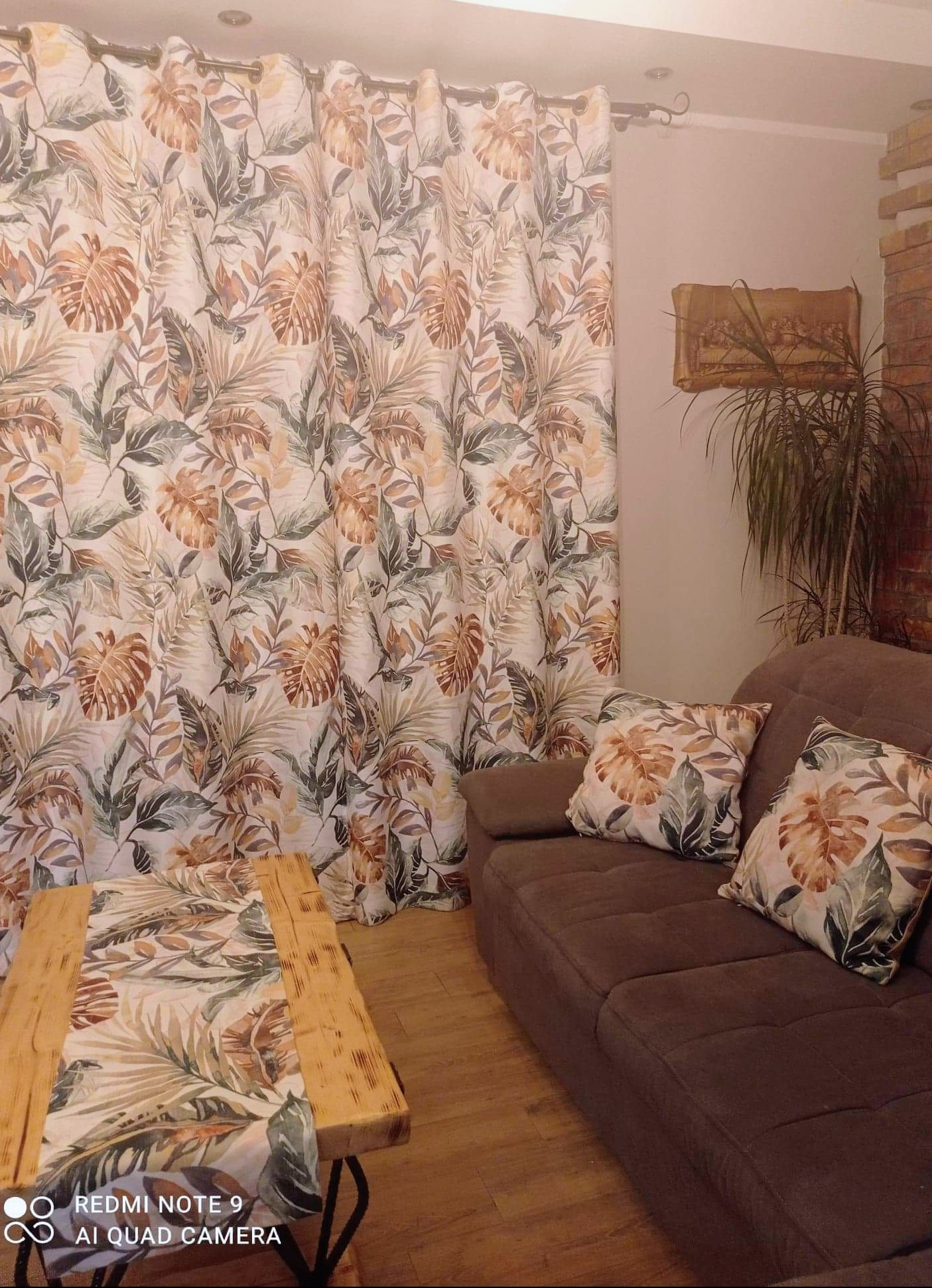 Ustawione zasłony w tropikalny wzór, dopasowane do poduszek na brązowej sofie i bieżnika na drewnianym stoliku kawowym. Całość w przytulnym wnętrzu.