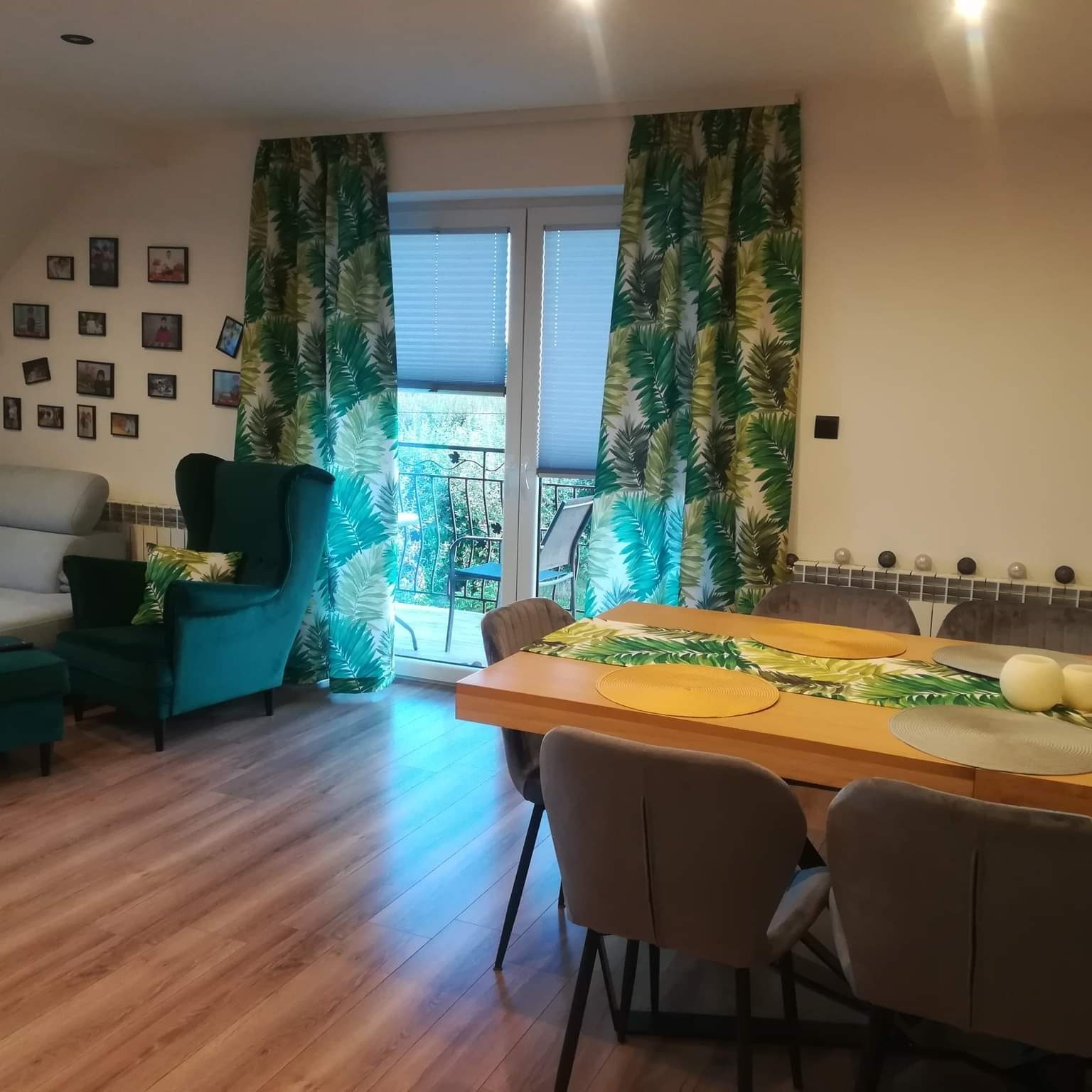 Salon z drewnianą podłogą i stołem. Na oknie zasłony w tropikalne liście, pasujące do poduszki na fotelu. Zdjęcia na ścianie dodają osobistego charakteru.