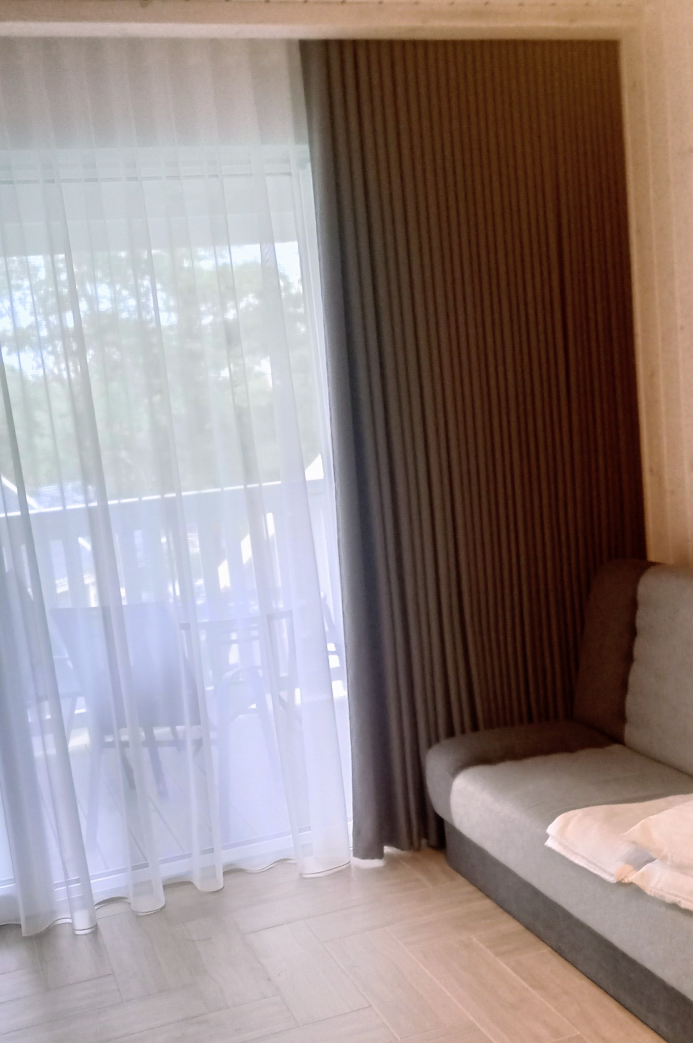 Jasne, zwiewne firany i ciężkie, brązowe zasłony w pokoju z szarą sofą. Widok z okna na balkon i drzewa. Kontrast faktur i kolorów.