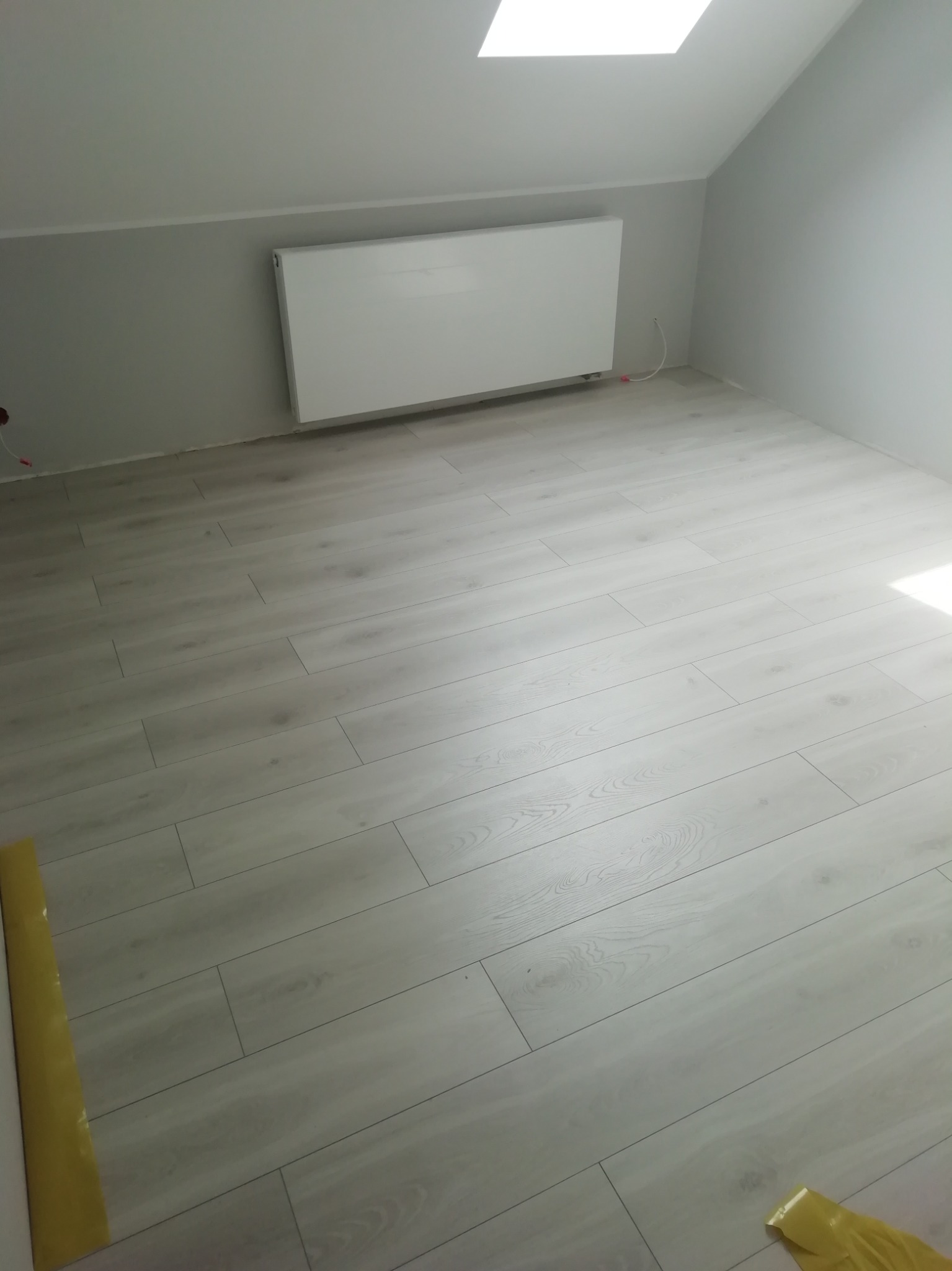 Jasnoszara podłoga z paneli ułożona w pokoju na poddaszu, widoczny biały grzejnik na szarej ścianie i fragment żółtej folii zabezpieczającej przy ścianie.