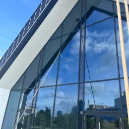 Czyszczenie szklanej elewacji budynku biurowego za pomocą lancy teleskopowej; w tle widoczne odbicie nieba i chmur.