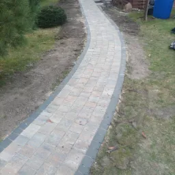 Zakrzywiona ścieżka z jasnoszarej kostki brukowej z ciemnoszarą obwódką prowadząca przez trawnik w kierunku drewnianej altany i niebieskiego pojemnika.