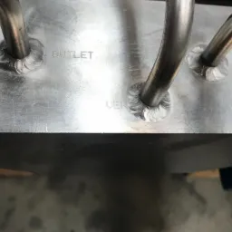 Szczegółowe ujęcie spawów łączących metalowe rury z aluminiową płytą, widoczne napisy 'OUTLET' i 'VEN'.