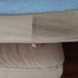 Fragment beżowej skórzanej kanapy z widoczną różnicą w czystości: po lewej stronie wyczyszczona, po prawej zabrudzona. Na górze leży niebieski koc. Widoczny zamek błyskawiczny i drewniana nóżka.