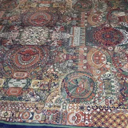 Wielobarwny dywan z geometrycznymi wzorami, głównie w odcieniach czerwieni, zieleni i brązu, z motywami okręgów i kwadratów, ukazany z perspektywy z góry.