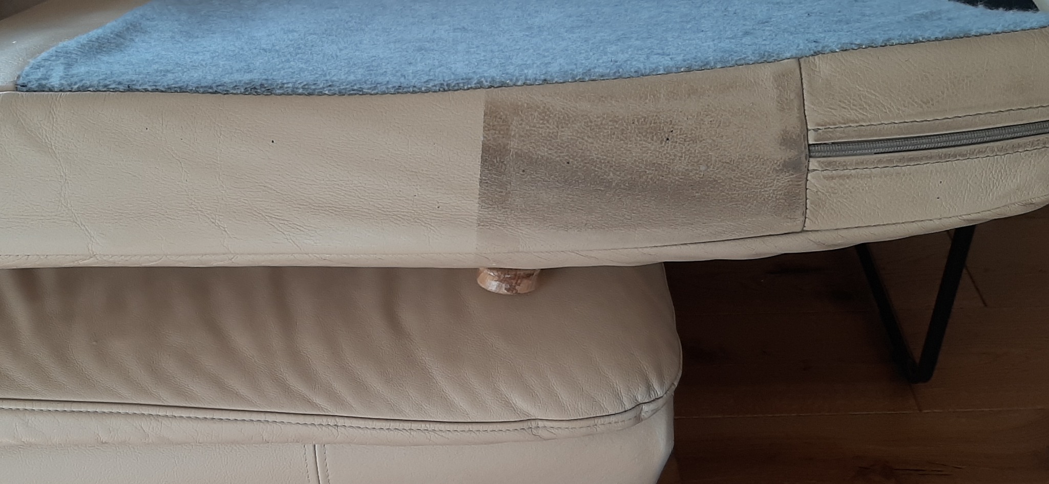 Fragment beżowej skórzanej kanapy z widoczną różnicą w czystości: po lewej stronie wyczyszczona, po prawej zabrudzona. Na górze leży niebieski koc. Widoczny zamek błyskawiczny i drewniana nóżka.