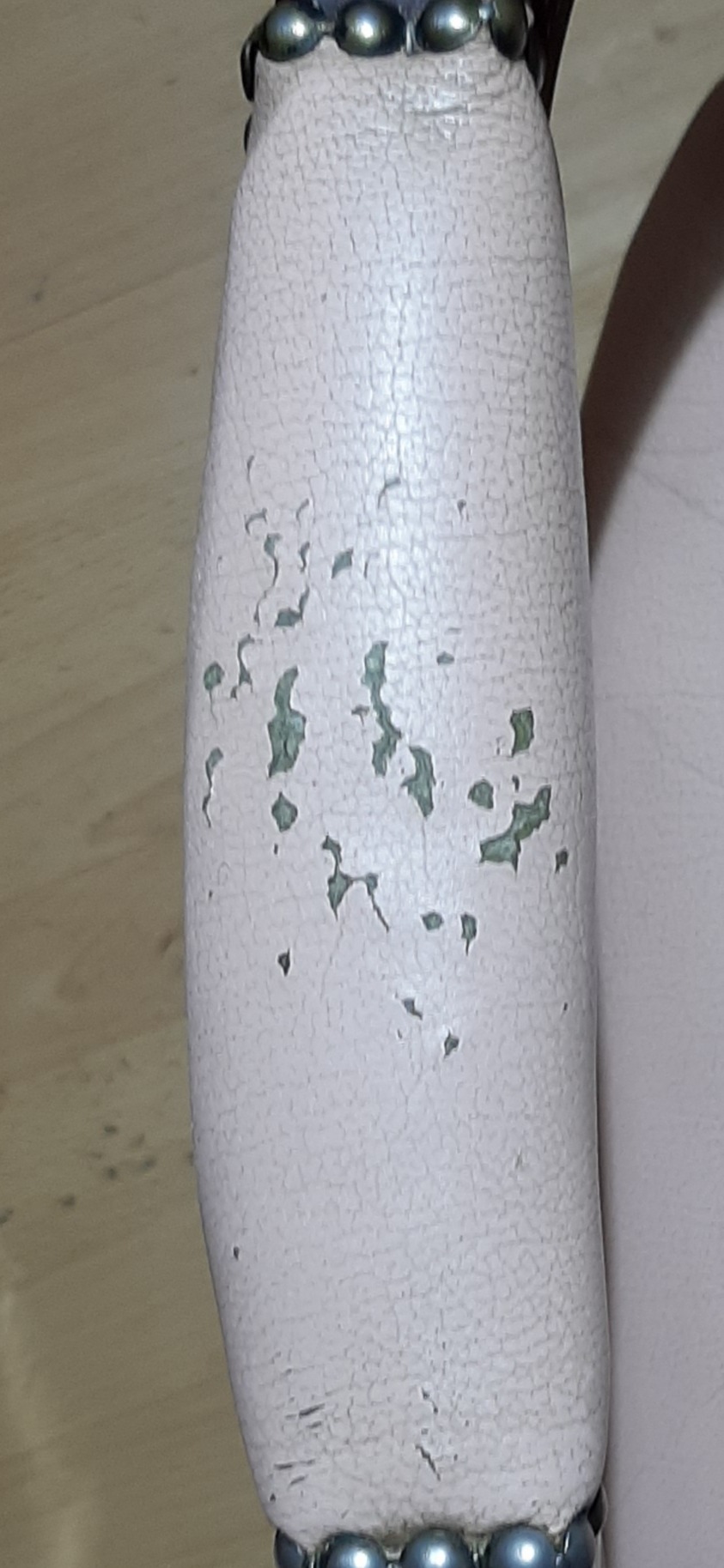 Podłokietnik fotela obity jasnoróżową skórą ekologiczną z widocznymi pęknięciami i odpryskami farby, wykończony ozdobnymi srebrnymi ćwiekami.