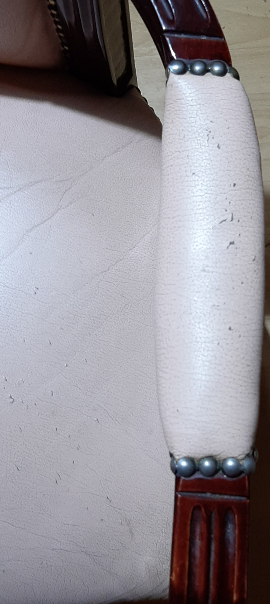 Fragment beżowego fotela z drewnianym, lakierowanym podłokietnikiem zdobionym srebrnymi nitami, widoczne drobne uszkodzenia tapicerki.