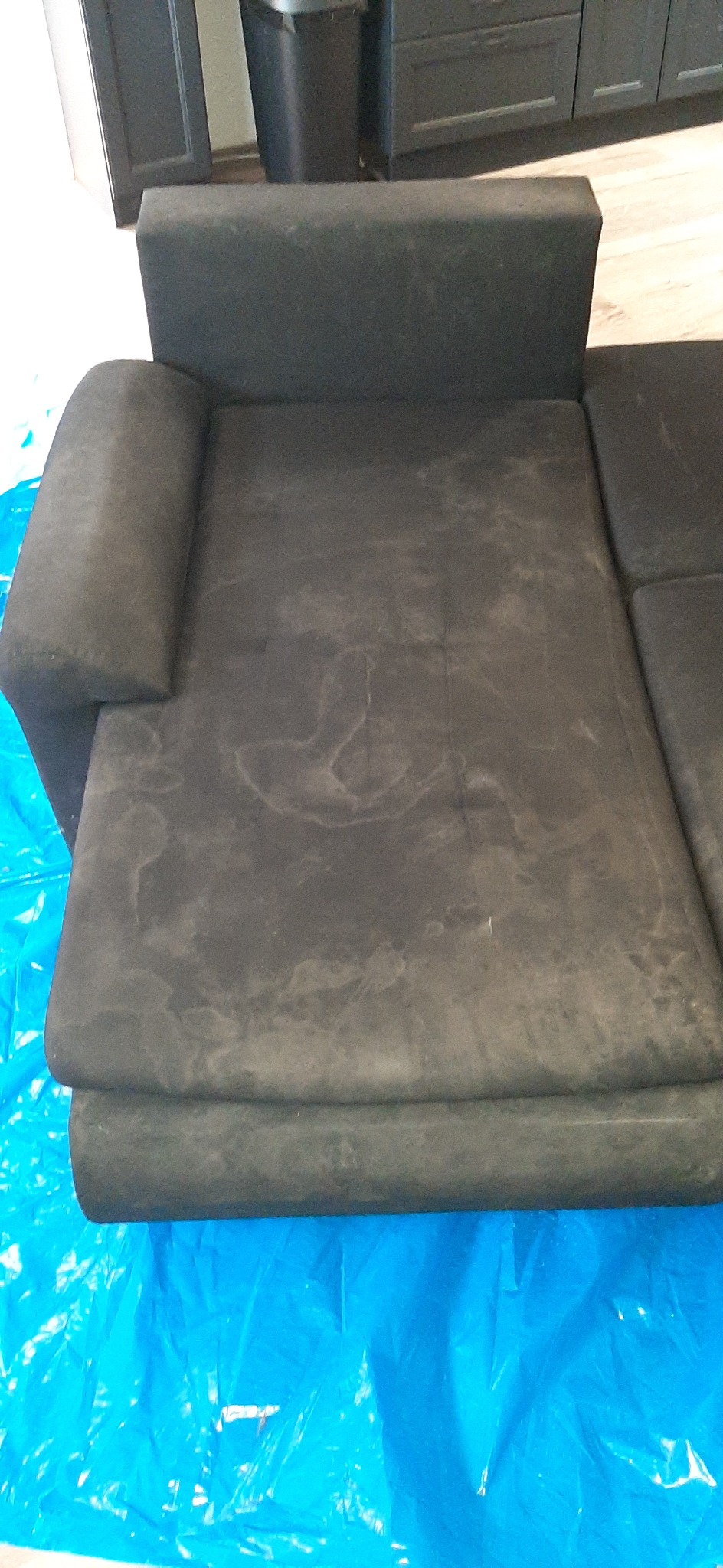 Ciemnoszara, tapicerowana kanapa z widocznymi plamami na siedzisku, zabezpieczona niebieską folią ochronną podczas czyszczenia.