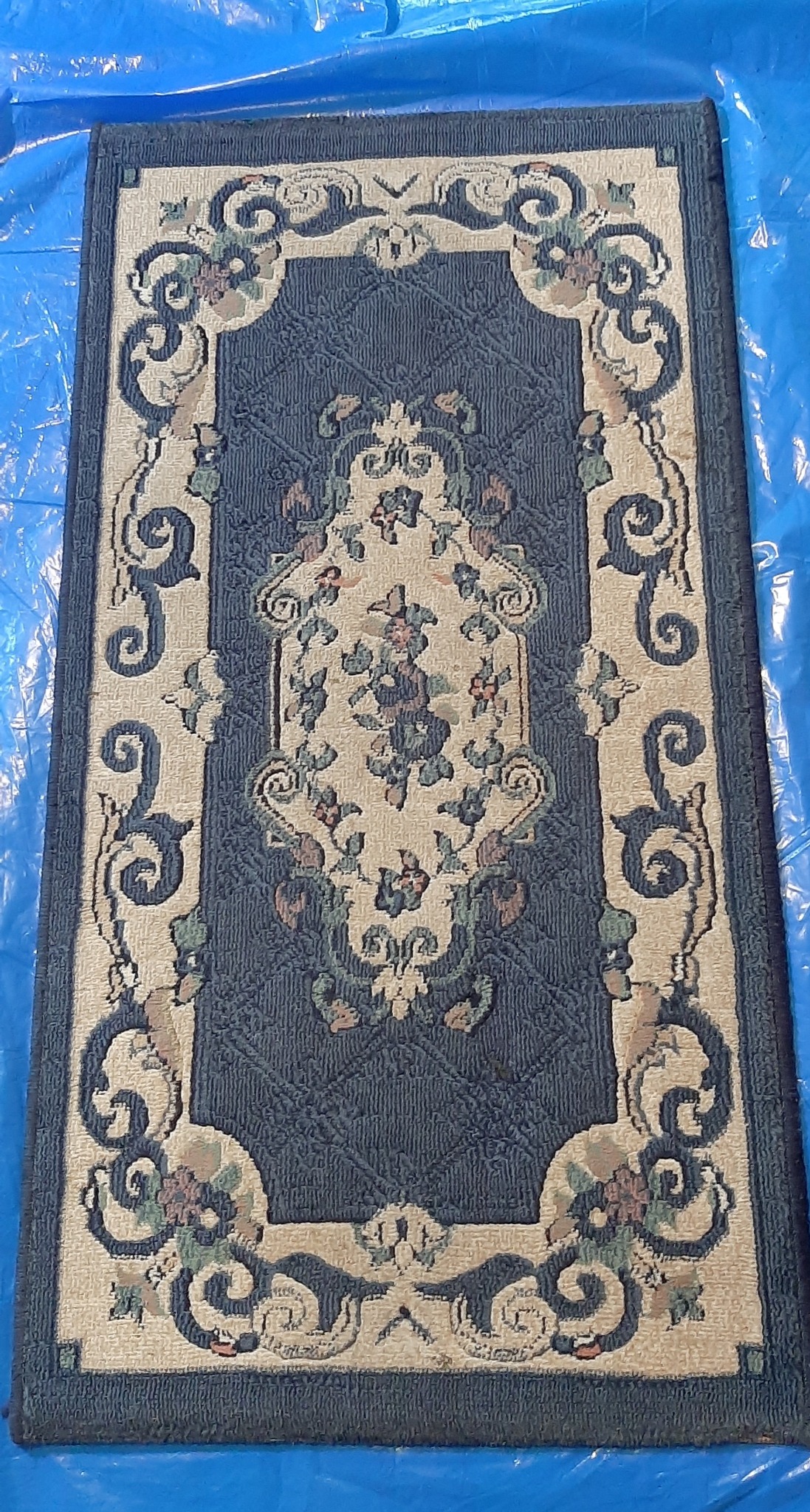 Prostokątny dywan z granatowym tłem i kremowo-beżowym obramowaniem z motywem kwiatowym, leżący na niebieskiej folii.