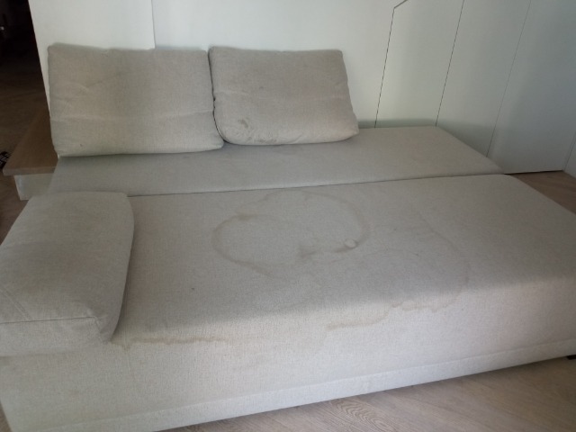 Jasna kanapa z widocznymi plamami na siedzisku i poduszkach, wymagająca czyszczenia tapicerki.