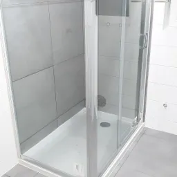 Kabina prysznicowa z przesuwnymi drzwiami, obłożona szarymi, kwadratowymi płytkami na ścianie i podłodze, z widocznym odpływem liniowym.