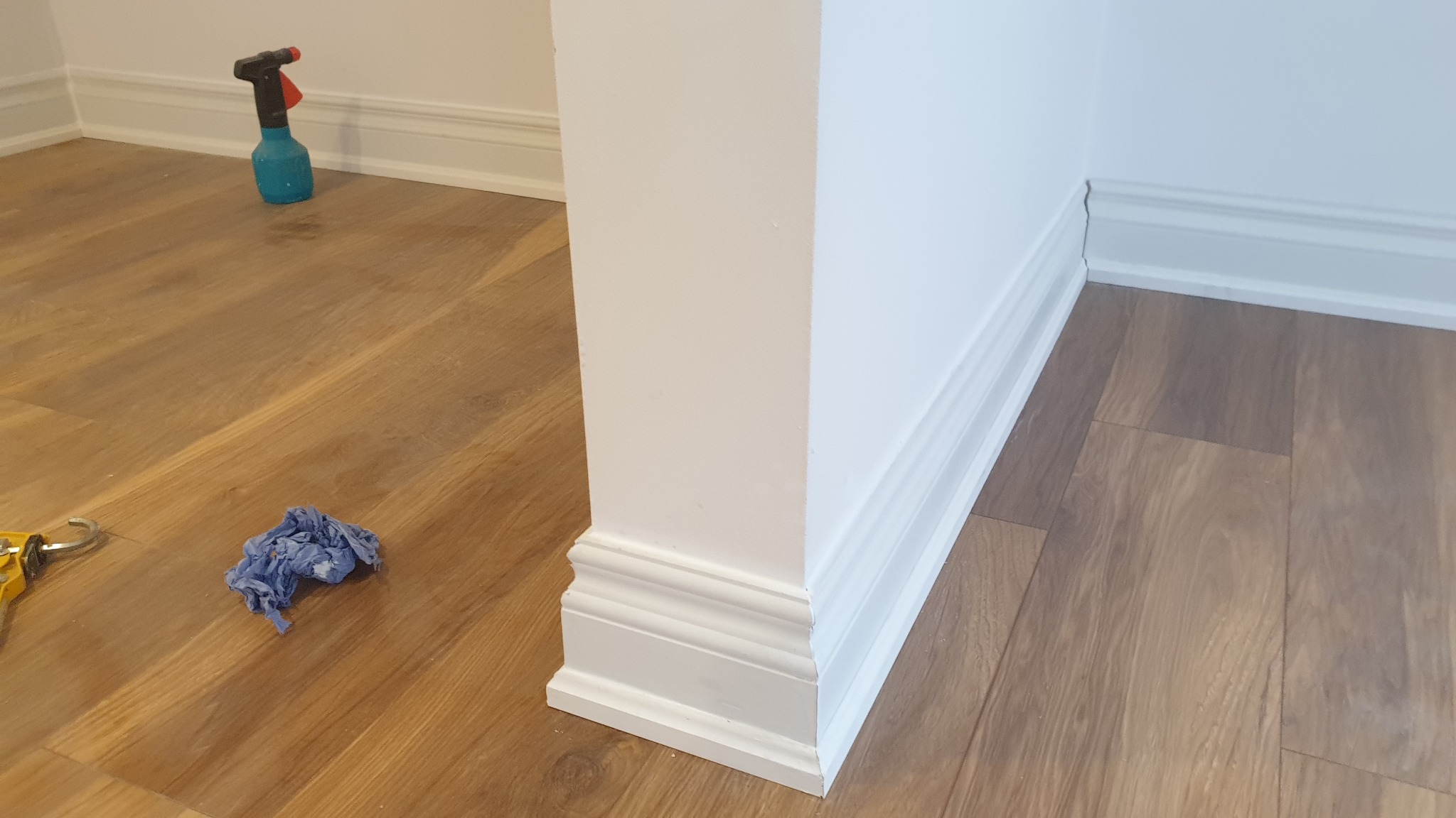 Świeżo pomalowane białe listwy przypodłogowe i narożnik ściany na tle podłogi z paneli, widoczny niebieski spryskiwacz i ścierka.