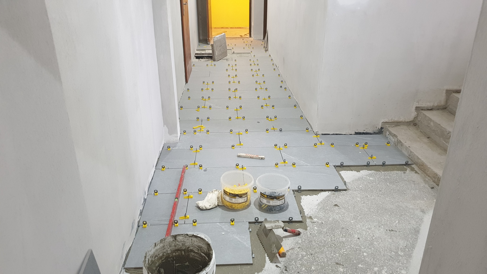 Korytarz w trakcie układania szarych płytek podłogowych z użyciem systemu poziomowania płytek, widoczne narzędzia glazurnicze, wiaderko i pojemniki z klinami.