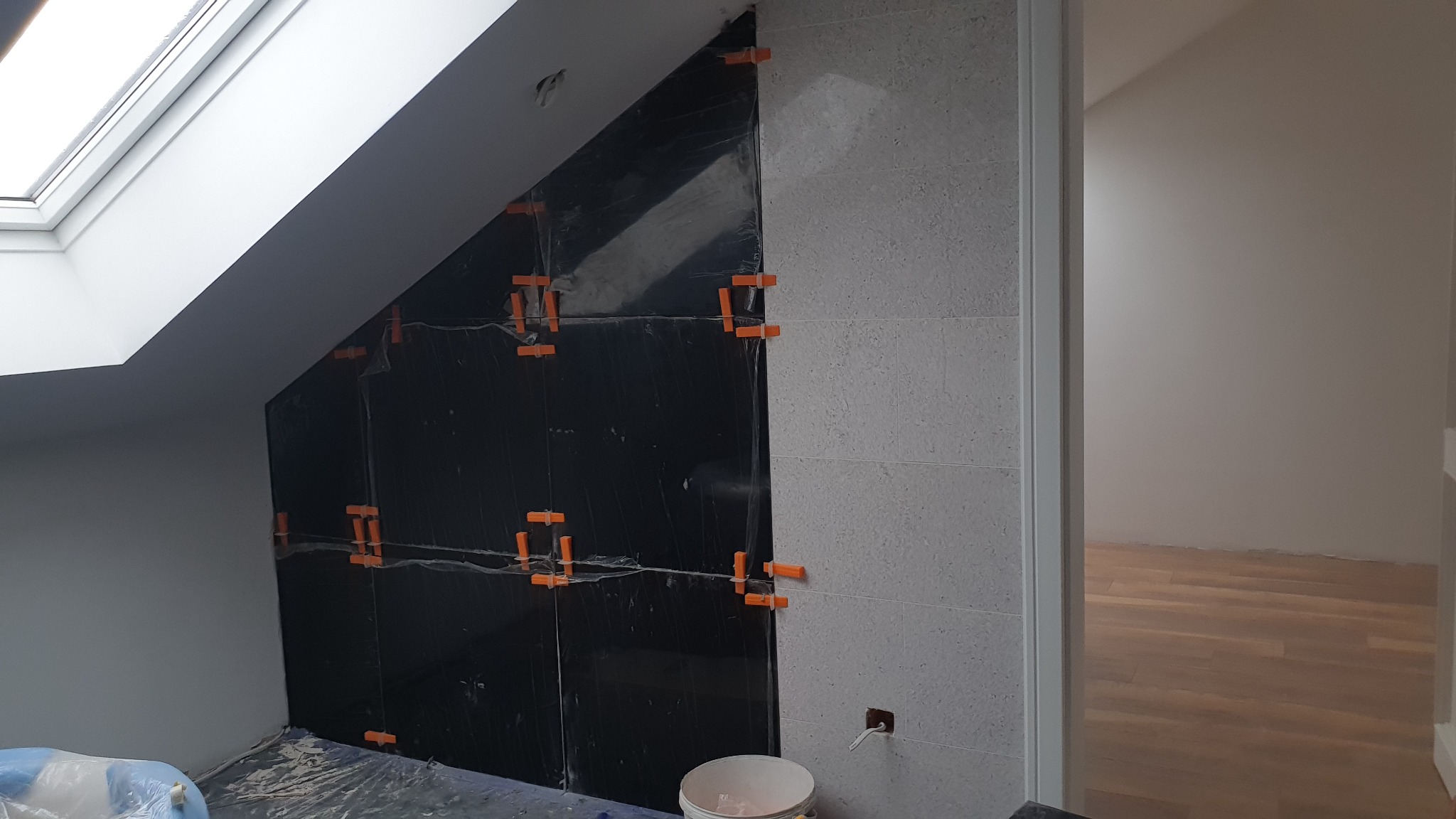 Ściana w trakcie wykańczania z częściowo ułożonymi czarnymi i szarymi płytkami, zabezpieczona folią, widoczne pomarańczowe krzyżyki dystansowe i wiadro z zaprawą.