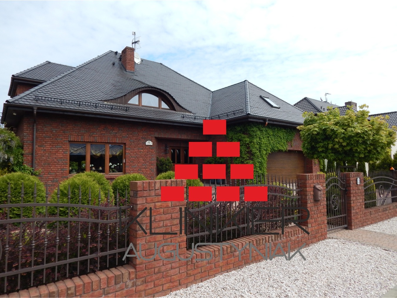 Dwukondygnacyjny dom z czerwonej cegły z ciemnoszarą dachówką, ogrodzony niskim, ceglanym murkiem z metalowym, ozdobnym płotem, częściowo porośnięty bluszczem.