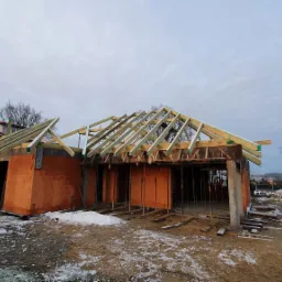 Budowa domu z czerwonej cegły: surowy stan z drewnianą konstrukcją dachu w trakcie montażu, widoczne deskowanie i elementy wsporcze, ziemia pokryta śniegiem.