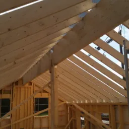 Konstrukcja dachu z drewnianych belek i metalowych łączników, widok z wnętrza budynku w trakcie budowy, z widocznym rusztowaniem i niebieskim niebem w tle.