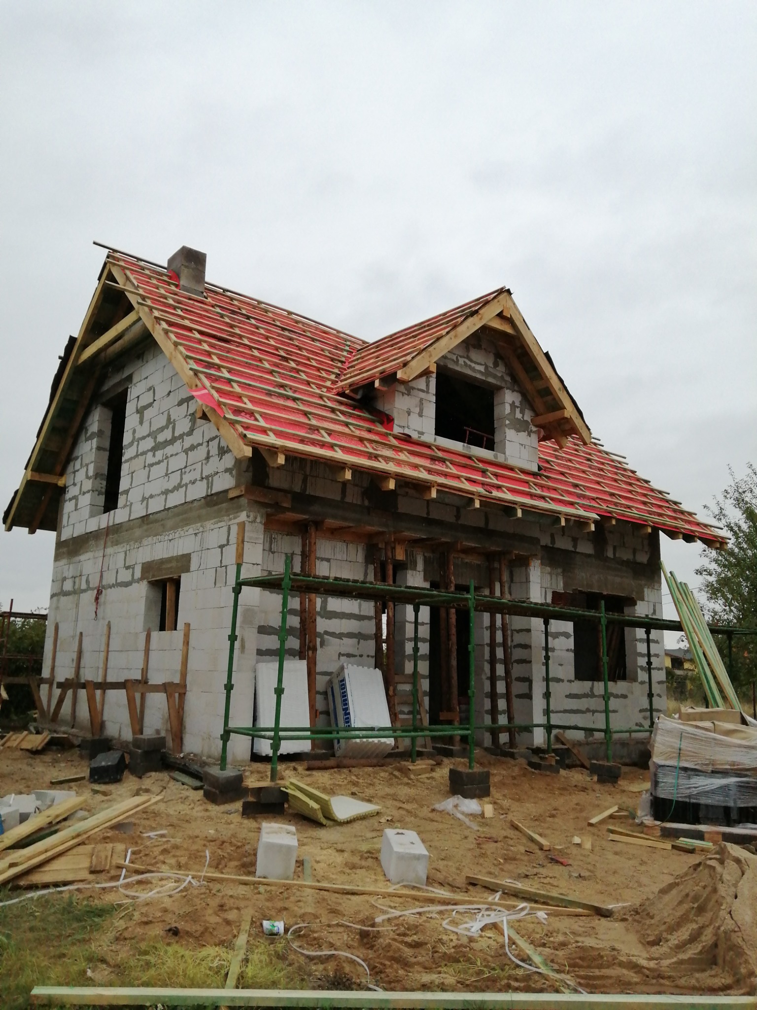 Dom murowany w trakcie budowy, widoczne ściany z bloczków, konstrukcja dachu z czerwoną folią, rusztowania i materiały budowlane na placu budowy.