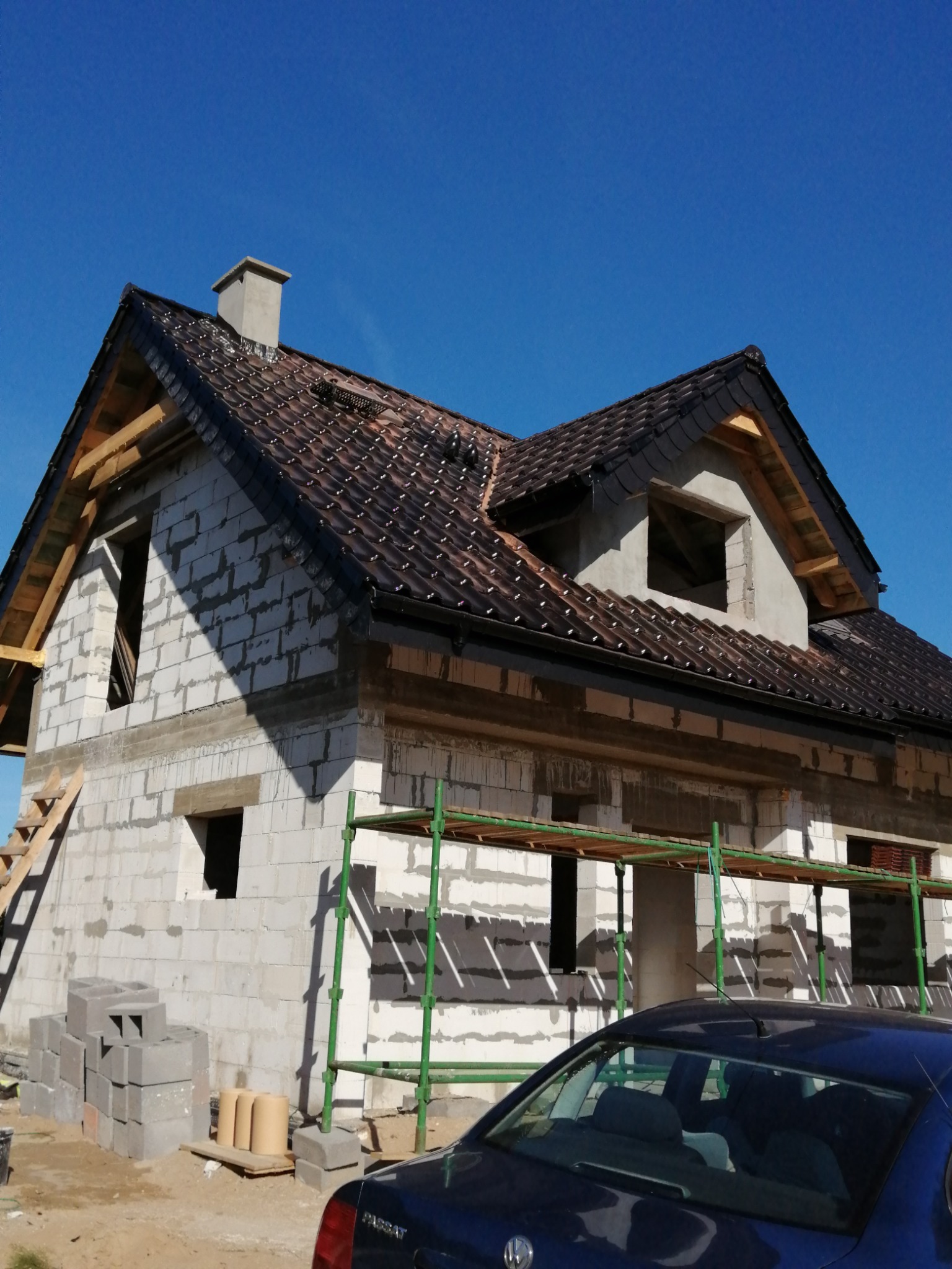 Niedokończony dom murowany z brązowym dachem z dachówki, widoczne rusztowanie, okna bez ram, oraz stos pustaków i zaparkowany samochód osobowy.