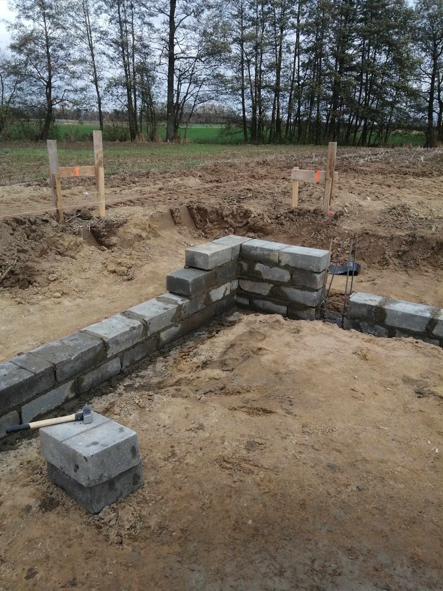Wznoszenie fundamentów z bloczków betonowych na placu budowy, widoczne drewniane znaczniki, w tle drzewa i pole.