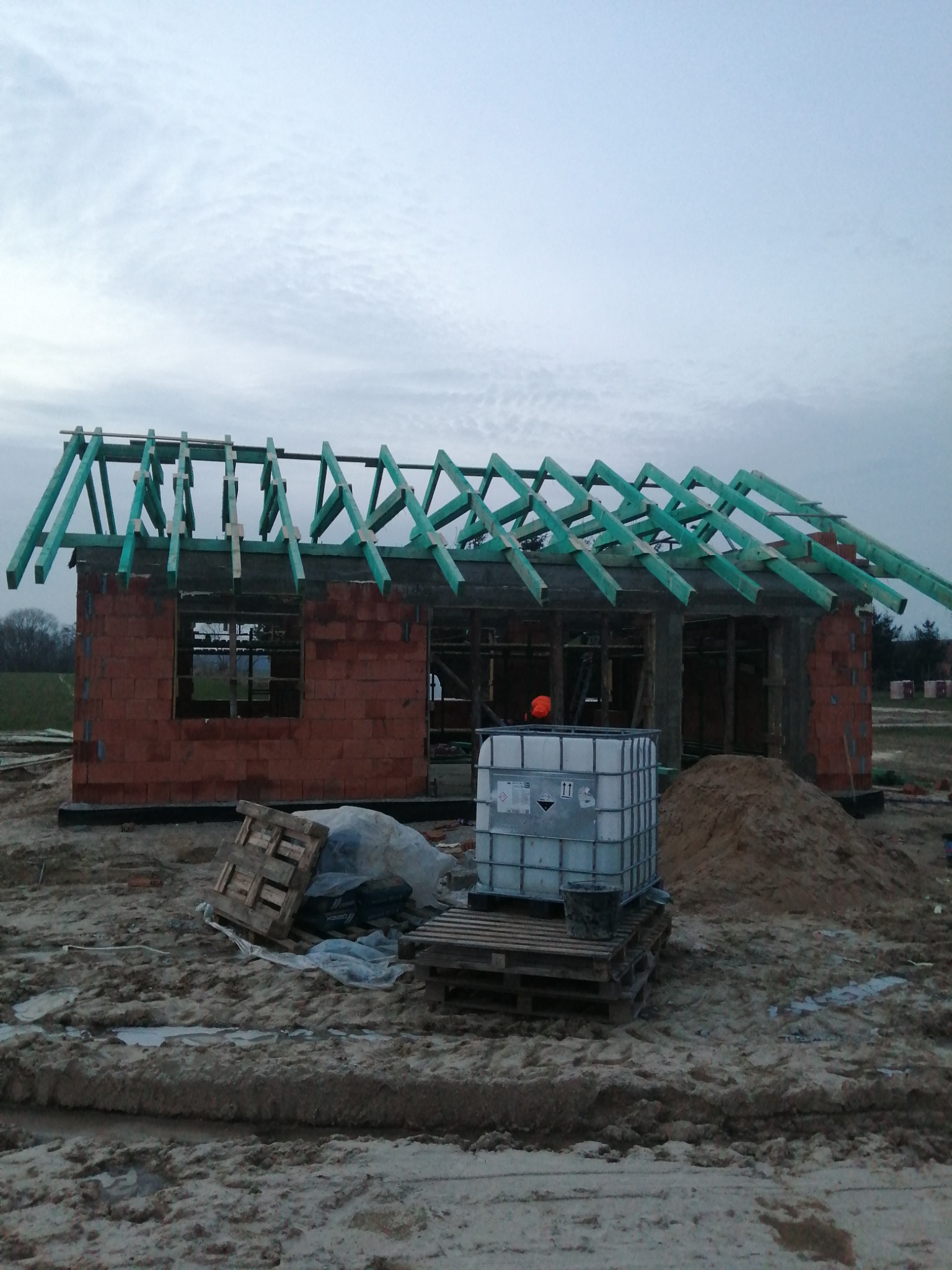 Budowa domu jednorodzinnego: surowy stan z czerwonej cegły z zielonymi krokwiami dachowymi, widoczny zbiornik na wodę i materiały budowlane na placu budowy.