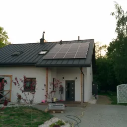 Dom jednorodzinny z ciemnym dachem, na którym zamontowano panele słoneczne; widoczne okna dachowe, komin z metalową osłoną, osoba stojąca na tarasie oraz elementy zagospodarowania terenu wokół...