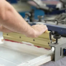 Sitodruk: ręczne nanoszenie farby na materiał za pomocą rakli w warsztacie, widoczny fragment maszyny i dłonie pracownika z zegarkiem.