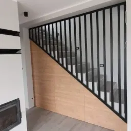 Nowoczesne schody z czarną metalową balustradą z pionowych prętów i obudową z paneli drewnopodobnych, widok z dołu na jasne stopnie schodów.