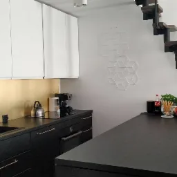 Nowoczesna kuchnia w Gdyni z czarnymi szafkami i złotym panelem nad blatem, minimalistyczna dekoracja ścienna i schody na antresolę z czarną balustradą.