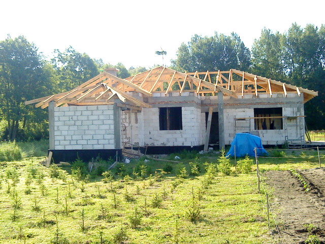 Surowy dom jednorodzinny w trakcie budowy z widoczną drewnianą konstrukcją więźby dachowej, bloczki z betonu komórkowego i rozciągnięta niebieska plandeka.
