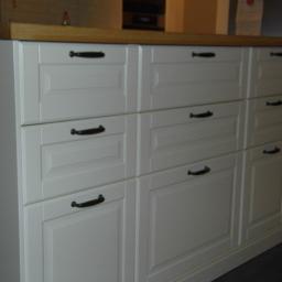 KRISMAL Krzysztof Bąkowski - Białe szafki kuchenne z ciemnymi uchwytami i drewnianym blatem, widoczne z bliska, z fragmentem podłogi w tle.