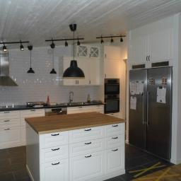 KRISMAL Krzysztof Bąkowski - Jasna kuchnia z białymi szafkami, drewnianym blatem wyspy, czarnymi uchwytami, metalowym okapem i oświetleniem szynowym z wiszącymi lampami.