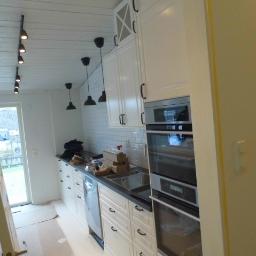 KRISMAL Krzysztof Bąkowski - Wnętrze kuchni w trakcie montażu: białe szafki z czarnymi uchwytami, zabudowany piekarnik, białe płytki na ścianie, sufit z drewnianą boazerią i czarnymi reflektorami szynowymi, widoczne kartony...