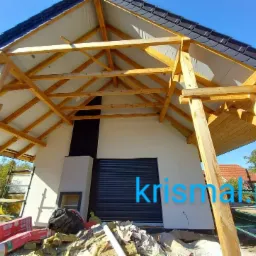 Konstrukcja drewniana dachu w trakcie budowy, widoczne krokwie, membrana dachowa, częściowo ułożona dachówka, biała ściana domu z czarną roletą okienną i kominem, oraz materiały budowlane na ziemi.