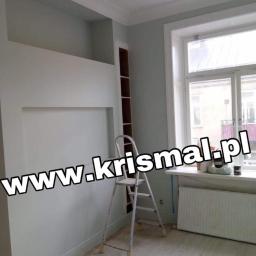 KRISMAL Krzysztof Bąkowski - Świeżo pomalowane wnętrze pokoju z wnękami w ścianie, białą drabinką, grzejnikiem pod oknem i jasną podłogą z desek. Ściany w odcieniach szarości i bieli. Widoczny fragment regału w zabudowie.