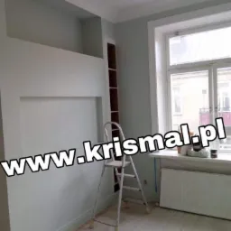 Świeżo pomalowane wnętrze pokoju z wnękami w ścianie, białą drabinką, grzejnikiem pod oknem i jasną podłogą z desek. Ściany w odcieniach szarości i bieli. Widoczny fragment regału w zabudowie.