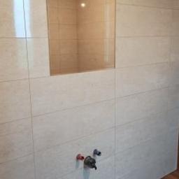 KRISMAL Krzysztof Bąkowski - Ściana łazienki wyłożona jasnymi, beżowymi płytkami, z wnęką wykończoną identycznymi płytkami. Widoczne przyłącza wodne z zaworami zamontowane na ścianie. Podłoga wyłożona panelami imitującymi drewno.