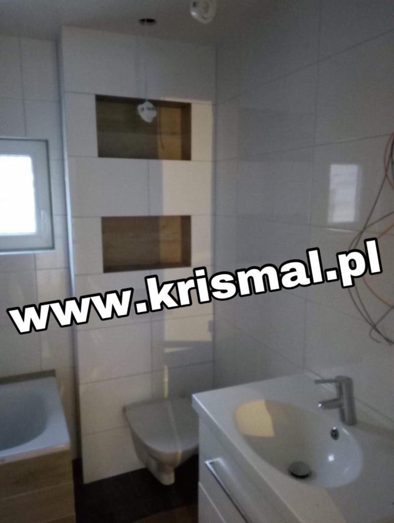 Biała łazienka z umywalką, toaletą podwieszaną i wnękami wykończonymi drewnem, widoczne kable elektryczne.