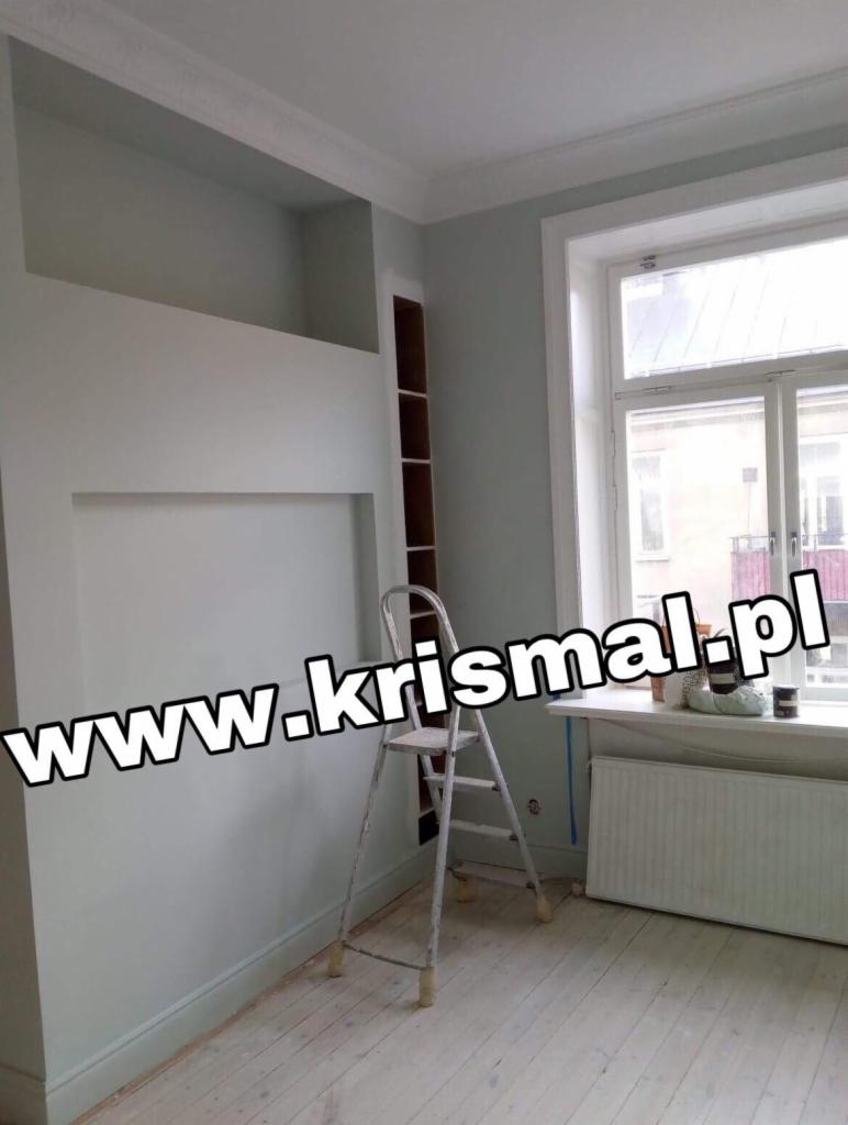 Świeżo pomalowane wnętrze pokoju z wnękami w ścianie, białą drabinką, grzejnikiem pod oknem i jasną podłogą z desek. Ściany w odcieniach szarości i bieli. Widoczny fragment regału w zabudowie.