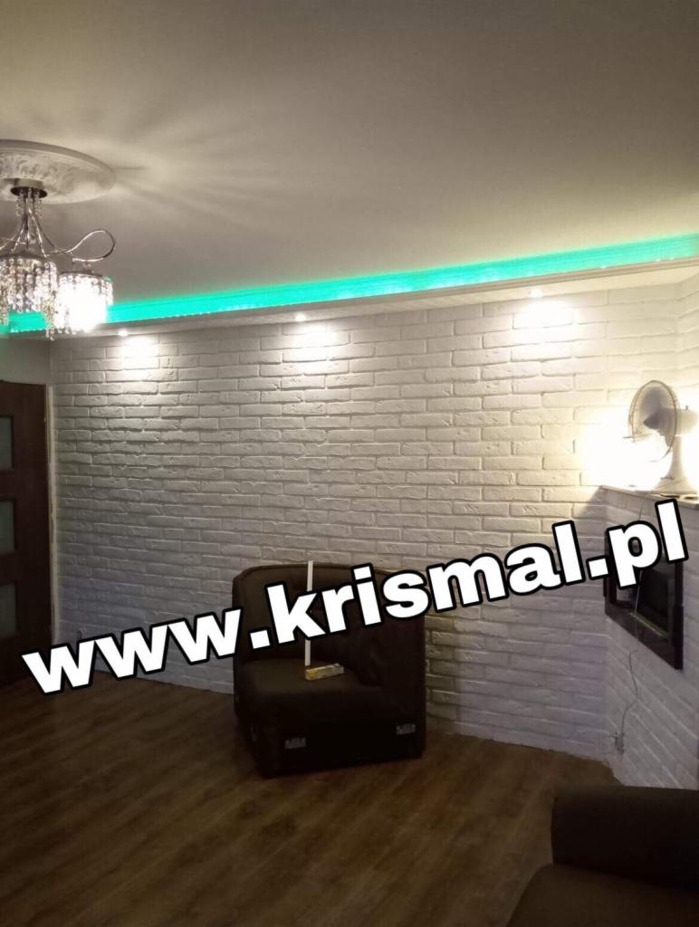 Biała ściana z cegły dekoracyjnej w salonie, podświetlona zielonym paskiem LED, z widocznym kominkiem i ciemnym fotelem. Na ścianie widoczny adres strony internetowej.