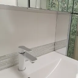 Biała umywalka z nowoczesną, chromowaną baterią, wykończona mozaiką w odcieniach szarości i białymi płytkami, odbijająca w lustrze fragment kabiny prysznicowej z motywem liści.