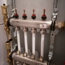 Rozdzielacz ogrzewania podłogowego z czterema pętlami, widoczne przepływomierze i rury doprowadzające, zamontowany na szarej ścianie.