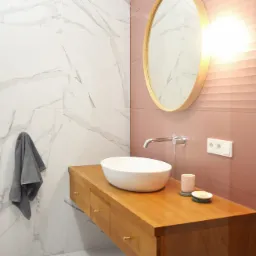 Nowoczesna łazienka z wiszącą szafką z jasnego drewna, umywalką nablatową i okrągłym lustrem w drewnianej ramie na tle różowej ściany z poziomymi żłobieniami. Marmurowe płytki na ścianach i podłodze.