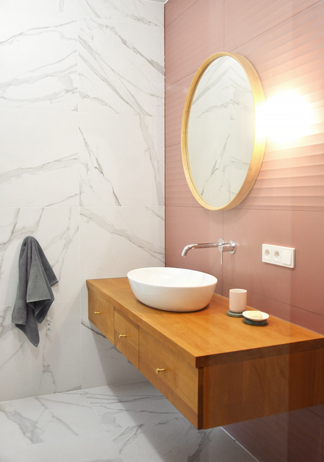 Nowoczesna łazienka z wiszącą szafką z jasnego drewna, umywalką nablatową i okrągłym lustrem w drewnianej ramie na tle różowej ściany z poziomymi żłobieniami. Marmurowe płytki na ścianach i podłodze.