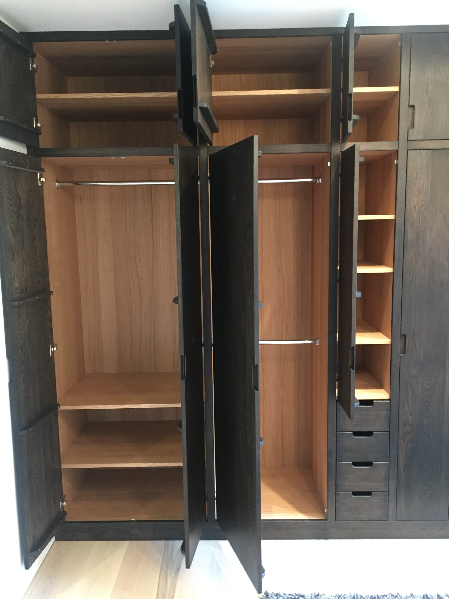 Wielodrzwiowa szafa z drewna w ciemnym odcieniu z widocznymi półkami, szufladami i drążkami na ubrania, niektóre drzwi otwarte, ukazujące jasne wnętrze.