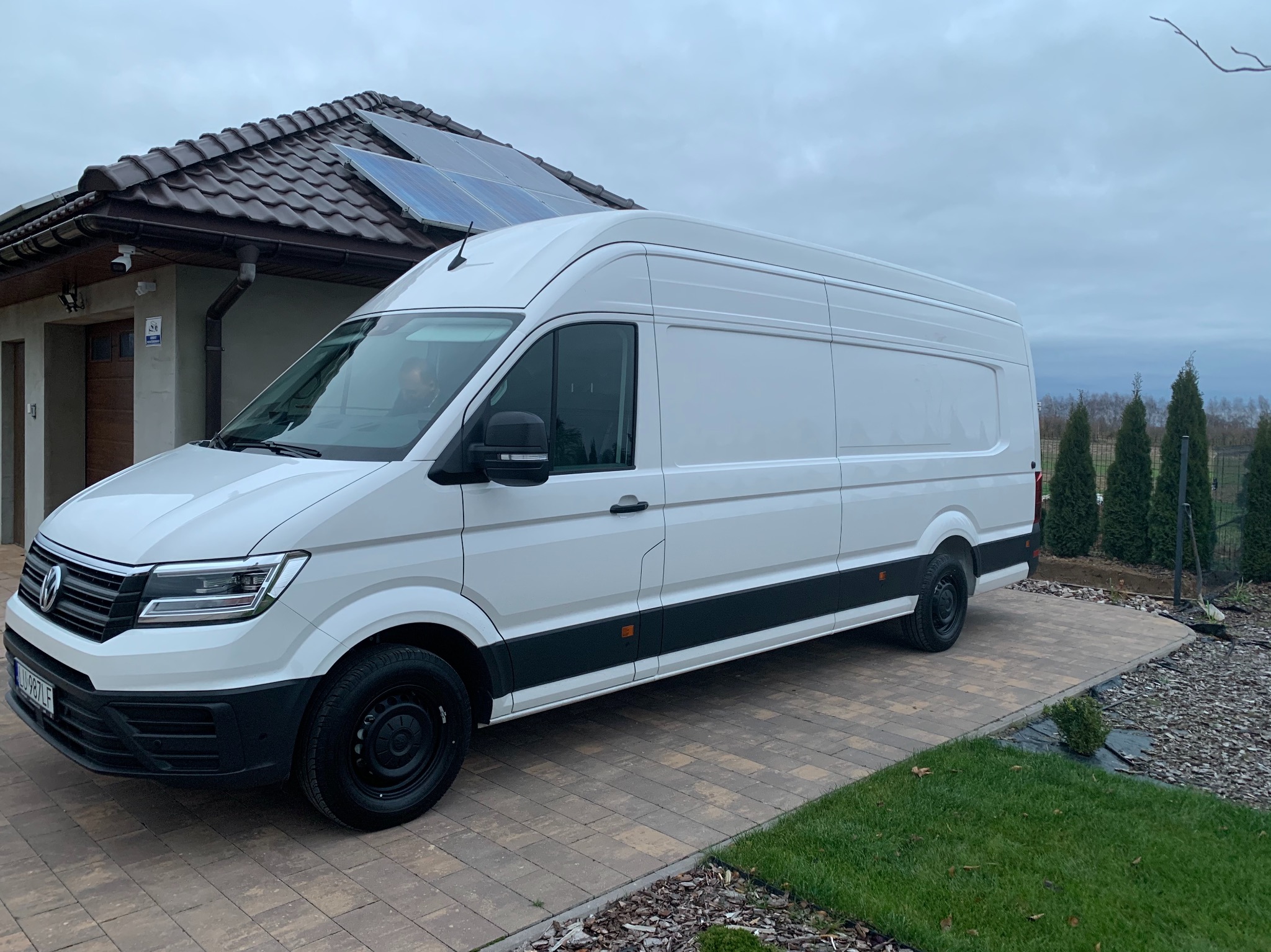 Biały, wysoki furgon marki Volkswagen, zaparkowany na brukowanym podjeździe przed domem z panelami słonecznymi na dachu, widok z boku.