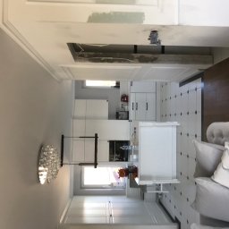 Grzegorz Osuch - Remont kuchni: widok na sufit z niedokończonym otworem, instalacja elektryczna widoczna, fragment pomalowanej ściany na próbę, w tle widoczna biała kuchnia z wyspą i kryształowym żyrandolem.